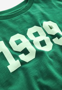 Grünes Baumwoll-T-Shirt mit großen, weißen Zahlen "1989", die auffällig in einer fett gedruckten Blockschrift bedruckt sind. Weiche Textur mit klassischem Rundhalsausschnitt.