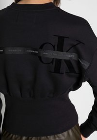 Sudadera negra con escote redondo, dobladillo acanalado y cintura fruncida. Presenta un gran estampado negro "ck" y una cinta de marca en la espalda.