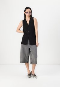 Schwarze ärmellose Weste mit V-Ausschnitt, ausgestattet mit Knöpfen und Taschen, kombiniert mit lockeren grauen Culottes und silbernen High Heels.