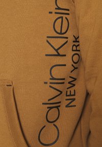 Sweatshirt marron en tissu doux, avec un grand texte noir "Calvin Klein NEW YORK" sur le côté et une poche kangourou.