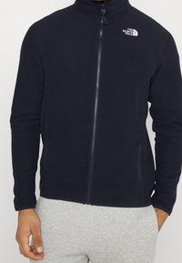 Svart fleecejacka med hög krage, full dragkedja och två sidofickor. Har en vit logotyp på den övre vänstra bröstområdet.