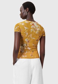 Geel bloemenpatroon top met korte mouwen en een aansluitend ontwerp, met een doorschijnende textuur en een ronde hals. Gemodeleerd met witte broek.
