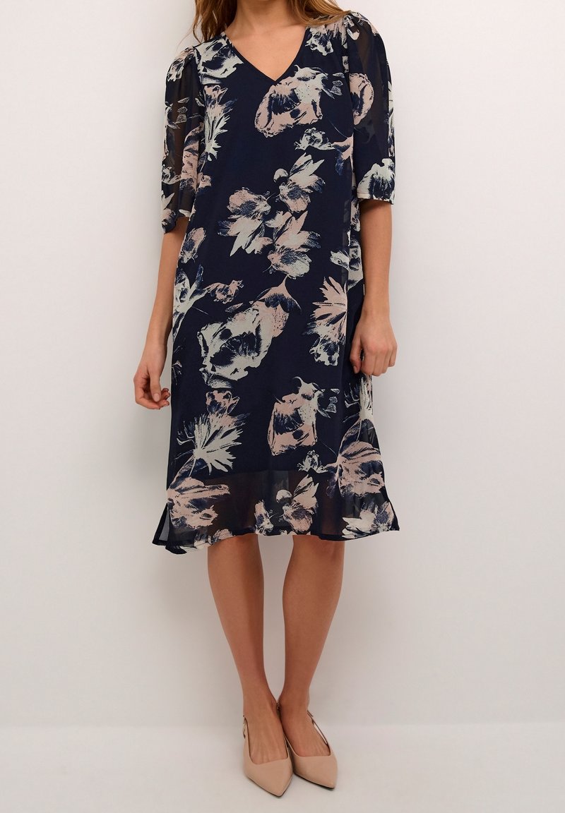 Femme portant une robe bleu marine longueur genou avec un imprimé floral clair, un col en V, des manches transparentes aux coudes et des talons pointus beige.