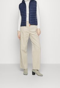 Gilet imbottito blu navy con cappuccio sopra una maglia bianca a maniche lunghe, abbinato a pantaloni beige a gamba larga e calzini alla caviglia verdi con tacco.