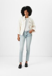 Off-white oversized jack, witte t-shirt, lichtblauwe jeans en zwarte instappers. Zwart krullend haar, staande tegen een witte achtergrond.