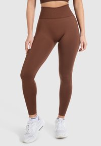 Braune, hochgeschnittene Leggings aus glattem Material, mit einer engen Passform und einem breiten Bund. Getragen mit weißen Turnschuhen.