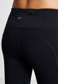 Mustat urheiluleggingsit, joissa on tiukka malli, litteät saumat ja pieni Nike-logo vyötäröllä sekä vetoketjullinen tasku takana.