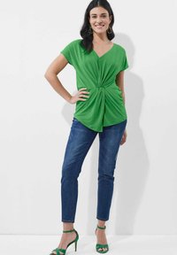 Grünes, kurzärmeliges Oberteil mit V-Ausschnitt und gedrehtem Vorderteil, kombiniert mit blauen Jeans und grünen, hochhackigen Sandalen.