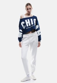 Sudadera recortada azul marino con letras blancas y texto "Universidad de California", combinada con pantalones jogger blancos y zapatillas blancas.