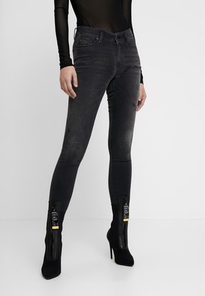 Mujer que lleva jeans ajustados negros deslavados con ligeros desgastes y botas de tacón alto negras hasta el tobillo con cremalleras frontales y etiquetas.