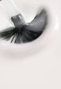 Brocha de maquillaje negra tocando un líquido gris, con las cerdas extendidas en abanico. Textura suave y cremosa rodea la brocha sobre un fondo claro.