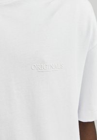 Balta medvilnė marškinėliai su įspaustu "ORIGINALS" logotipu ant krūtinės. Laisvas kirpimas, trumpomis rankovėmis ir švelniu, lygiu tekstūru.