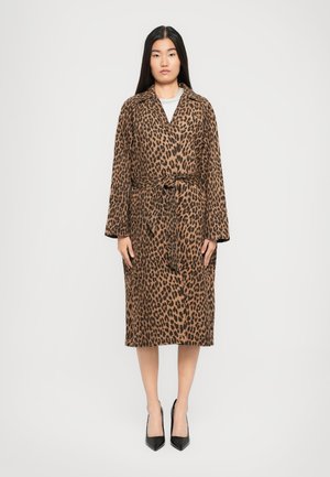 Žena stoji uspravno, odjevena u smeđi kaput sa leopard uzorkom do koljena i crne visoke potpetice na jednoličnoj pozadini.