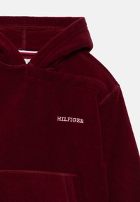 Haut à capuche en polaire bordeaux avec cordon de serrage, poche frontale et logo "HILFIGER" blanc. Texture douce et couleur unie avec un minimum de détails.