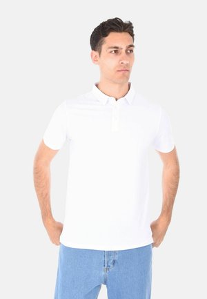 Uomo con capelli scuri che indossa una polo bianca a maniche corte e jeans azzurri chiaro, in piedi con le mani nelle tasche, con lo sguardo rivolto di lato.