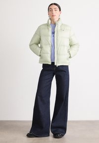 Ljusgrön pufferjacka med hög krage, framzipper och sidofickor, kombinerad med mörkblå vida jeans och svarta skor.