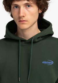 Grøn hætte sweatshirt med snøre, der har et blåt logo på venstre bryst. Lavet af blødt stof med en tekstureret overflade og kængurulomme.