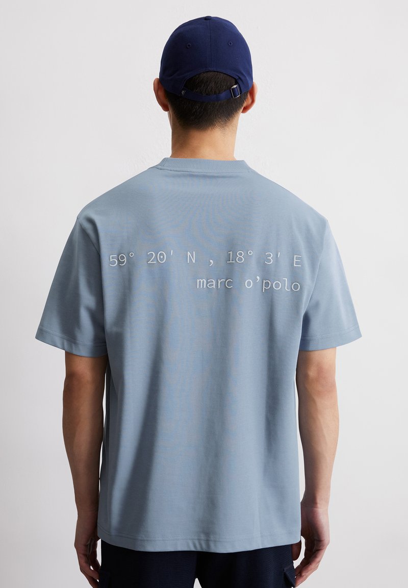 Jasnoniebieska bawełniana koszulka z okrągłym dekoltem, krótkimi rękawami i haftowanym napisem na plecach w białym kolorze: "59° 20'N, 18° 3'E marc o'polo."