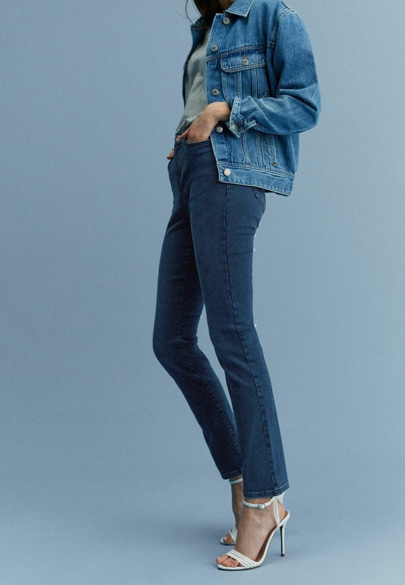 Chaqueta de denim en azul claro, camiseta gris ajustada, vaqueros ajustados azul oscuro y tacones blancos con tiras. Textura suave y diseño clásico en todo.