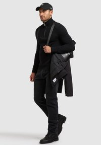Maglione nero con colletto zip, jeans neri e una giacca imbottita nera poggiata su un braccio. Berretto nero e stivali robusti neri completano l'outfit.