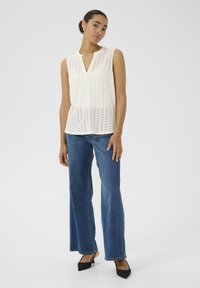 Blouse sans manches crème avec des détails en œillets, décolleté en V, associée à un jean en denim large bleu et des escarpins noirs à bout pointu.