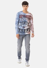 Langärmliges T-Shirt mit Batikmuster in Blau und Grau, versehen mit grafischem Druck, kombiniert mit ausgewaschenen grauen Jeans mit Knietaschen und Nähdetails.