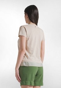 Camiseta de manga corta beige con cuello redondo, superpuesta sobre un dobladillo verde claro. Tejido suave con un pequeño acento rojo en la parte trasera.
