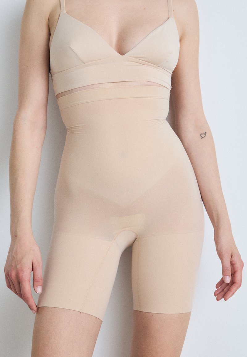 Beige high-waisted shapewear shorts met een gladde textuur en een aansluitend ontwerp, met een naaddetail langs de benen en een lengte tot halverwege de dij.