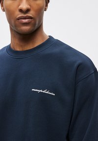 Tmavomodrá mikina s rebrovaným okrúhlym výstrihom a jemnou textúrou. Na hrudi je biela kurzívna výšivka. Jednoduchý a moderný dizajn.