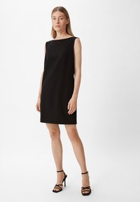 Schwarzes ärmelloses Kleid mit geradem Schnitt, ausgestattet mit einem Bootsausschnitt und glatter Textur. Kombiniert mit schwarzen Riemchen-High-Heels.