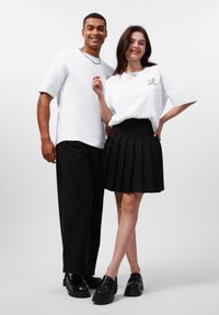 Bijele oversize majice na modelima, jedan u crnoj plisiranoj suknji, a drugi u crnim širokim hlačama, oboje nose crne cipele i lancasti ogrlicu.