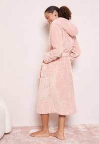 Ružová chlpová župan s kapucňou, s vlnitou textúrou, dlhými rukávmi a opaskom. Obsahuje bočné vrecká a siaha pod kolená.