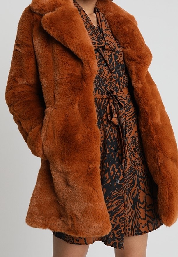 Puszysty, pomarańczowo-brązowy płaszcz z sztucznego futra z szerokim kołnierzem, długimi rękawami i luźnym krojem. Noszony na wzorzystej, ciemnej sukience.