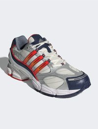 Weiße und marineblaue Sportschuhe mit roten Akzenten, strukturiertem Mesh-Obermaterial, dicker Sohle und weißen Schnürsenkeln vor einem hellgrauen Hintergrund.