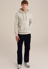 Bež kapuca s sprednjim žepom, kapuco s vrvico in rebrastimi manšetami, v kombinaciji z temno modrimi žametalnimi hlačami in sivimi supergami.