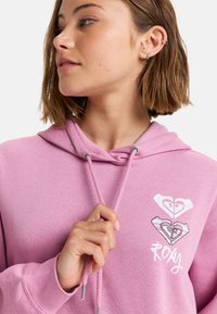 Růžová mikina s kapucí, která má přední kapsu a šňůrky na stažení. Grafický design se dvěma tvary srdce a názvem značky "Roxy" vytištěným bílou barvou.