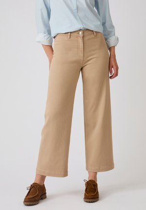 Woman wearing beige wide-leg pants with front button and brown lace-up shoes, standing against a plain light background.