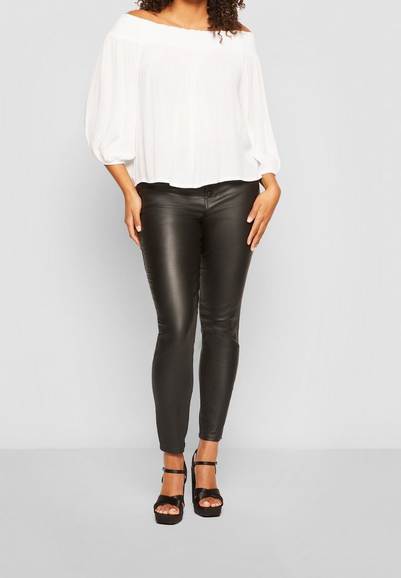 Femme portant un chemisier blanc épaules nues, un pantalon en cuir noir et des sandales noires à talons hauts, debout devant un fond uni.
