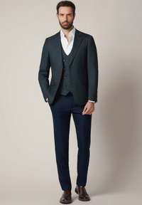 Mężczyzna w ciemnozielonej marynarce, dopasowanej kamizelce, białej koszuli, granatowych spodniach i brązowych skórzanych butach stoi na tle jednolitego beżowego tła.