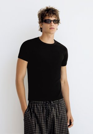 Jeune homme portant un t-shirt noir ajusté, des lunettes de soleil et un pantalon à cordon à carreaux, debout devant un fond blanc.