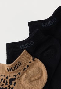 Drie paar sokken: één zwart met geribbelde textuur, één donkerblauw met logo, en één beige met zwart patroon en logo bij de rand.