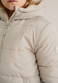Švarkas pūkų paltas šviesiai béže spalvos su minkštu pūkiniu gobtuvu. Yra įstrižai siuvinėtos linijos, priekinis užtrauktukas ir apvalus logotipo ženklelis ant rankovės.