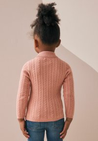 Rosa Strickpullover mit einem Zopfmuster, geripptem Kragen und langen Ärmeln, kombiniert mit blauen Jeans. Die Ansicht zeigt die Rückseite des Outfits.