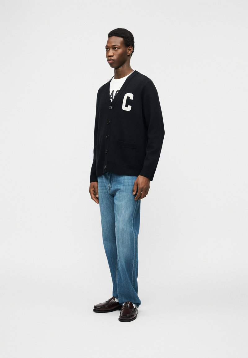 Homme debout portant un cardigan noir boutonné avec la lettre C, une chemise blanche, un jean bleu et des mocassins marron foncé sur fond blanc.