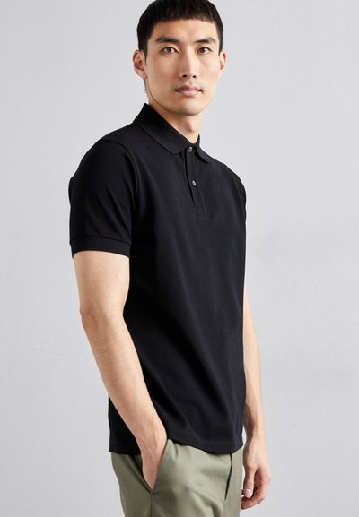 Polo noir en coton, avec une patte de boutonnage à deux boutons, des manches courtes et une coupe ajustée. Porté avec un pantalon vert clair.