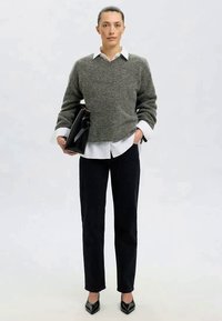Grauer Wollpullover über einem weißen Hemd mit Kragen, kombiniert mit schwarzen Hosen und schwarzen High Heels; hält eine schwarze Handtasche; minimalistisches Design.