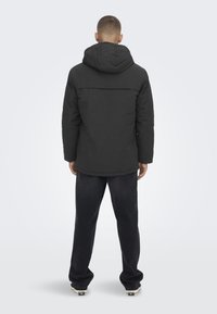 Chaqueta acolchada negra con capucha, con cierre frontal y corte recto, combinada con pantalones oscuros y zapatos casuales.