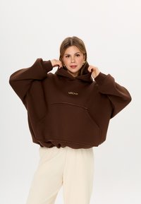 Felpa oversize marrone con tasca frontale e logo ricamato. Tessuto morbido con orlo e dettagli a coste. Abbinata a pantaloni chiari.