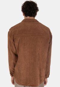 Camicia di corduroy marrone con righe verticali, maniche lunghe e dettaglio della spalla sul retro. Presenta una vestibilità morbida e un orlo curvo nella parte inferiore.