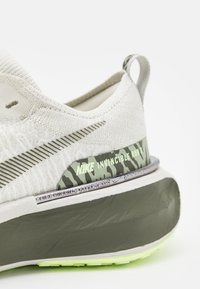 Gaiši pelēki Nike Invincible Run apavi ar teksturētu adītu augšdaļu, zaļiem akcentiem un biezām, mīkstinātām zolēm ar rakstainu vidusdaļu.